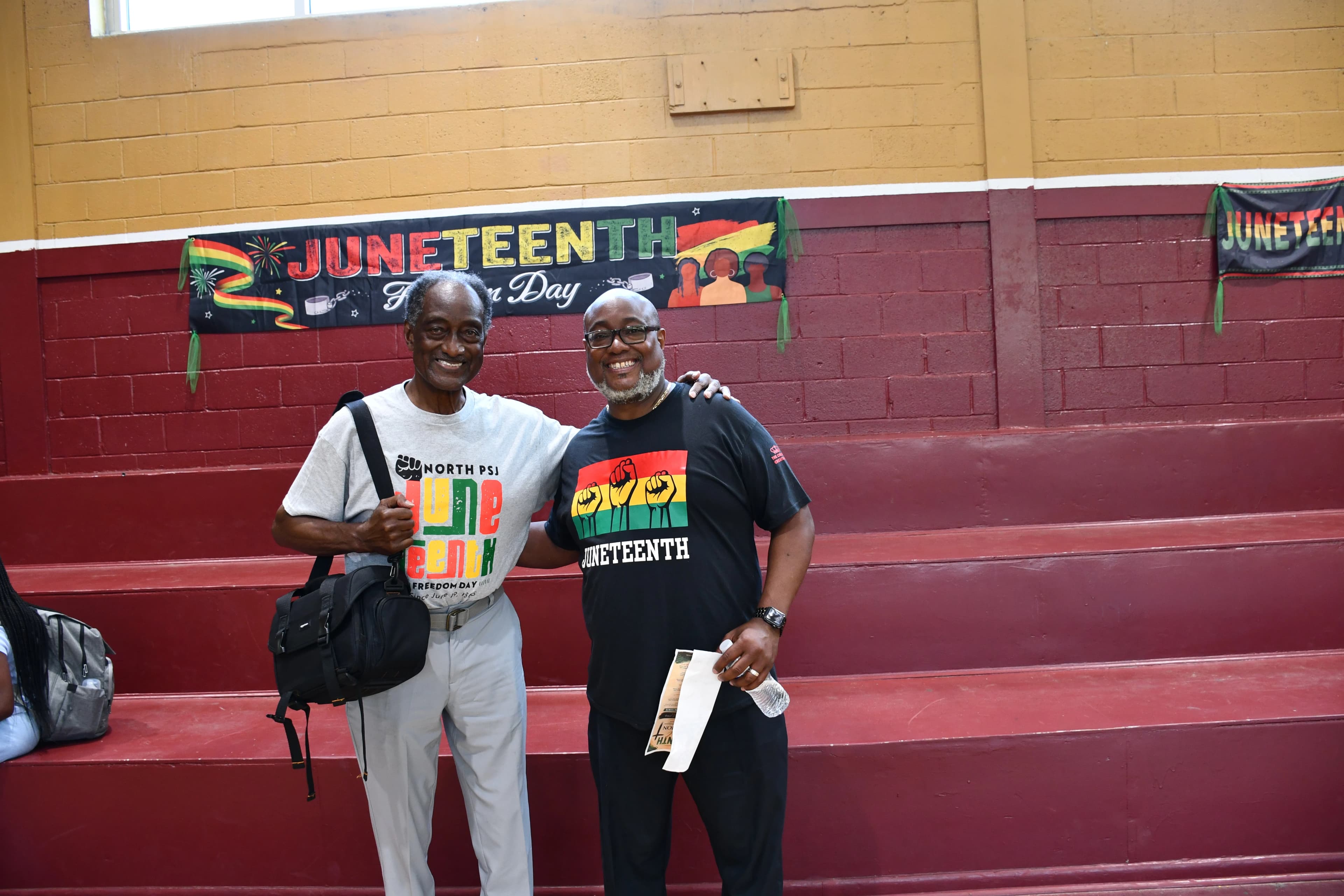 Two community members in Juneteenth t-shirts posing in front of a Juneteenth Freedom Day banner