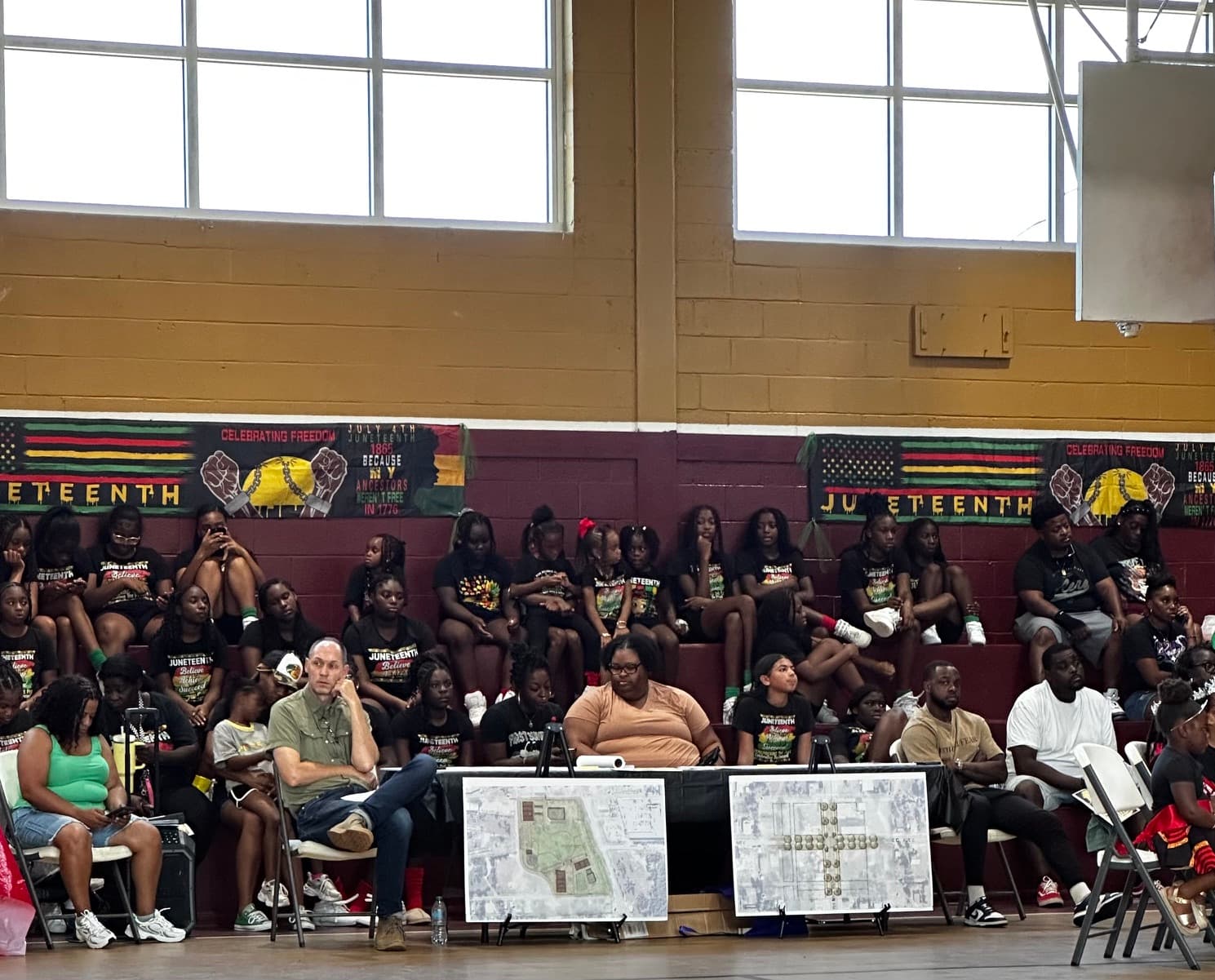 Attendees and youth in Juneteenth t-shirts in the bleachers with community planning maps on display